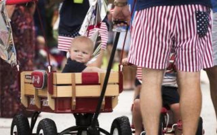 Child in wagon pushed by person in American flag shorts.